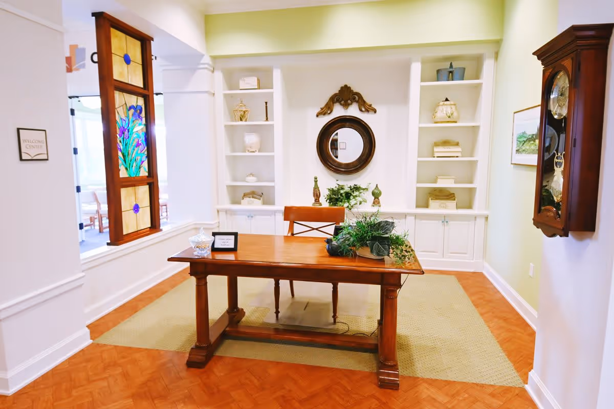 A warmly lit interior room featuring a wooden desk with a chair behind it, decorated with a plant and a small sign. Behind the desk is a built-in white shelving unit with decorative items and a round mirror with an ornate wooden frame. To the left, a stained glass panel with floral design hangs near a doorway labeled 'Welcome Center'. On the right wall, a wooden pendulum clock is mounted.
