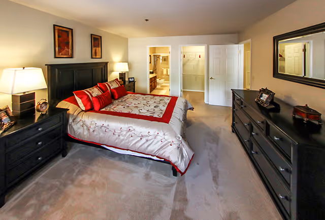 Spacious bedroom with a large bed dressed in red-accented bedding, dark wood dressers and nightstands, and an en-suite bathroom visible through open doors.
