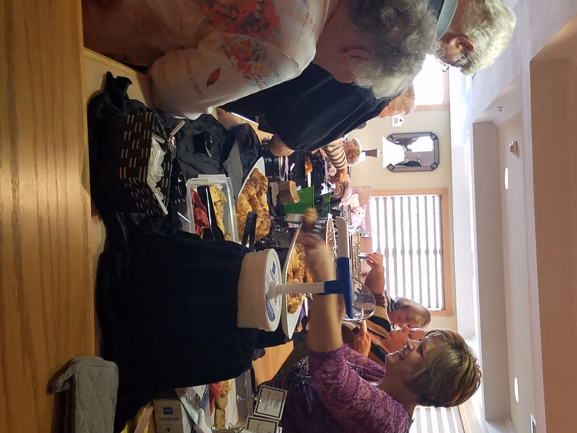A group of elderly women standing around a long table filled with various pastries and food items in a well-lit room with windows and a mirror on the wall.
