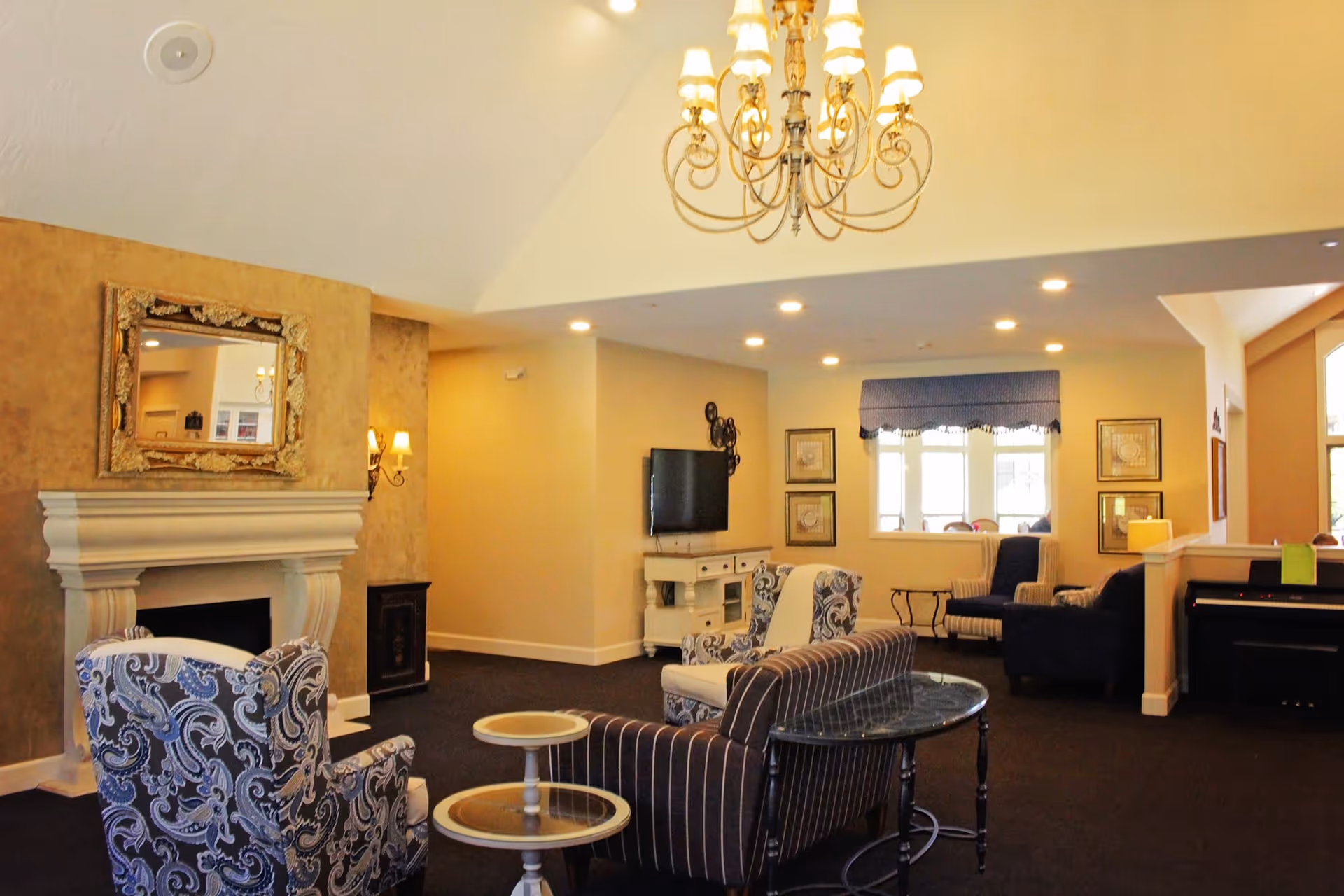 Bright furnished common living room with patterned armchairs, a fireplace, chandelier, TV and multiple seating areas.