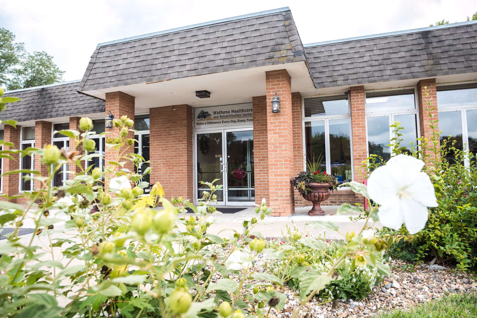 Exterior view of Wathena Healthcare and Rehabilitation Center showing the entrance with glass double doors, brick pillars, and a garden with green plants and a white flower in the foreground.
