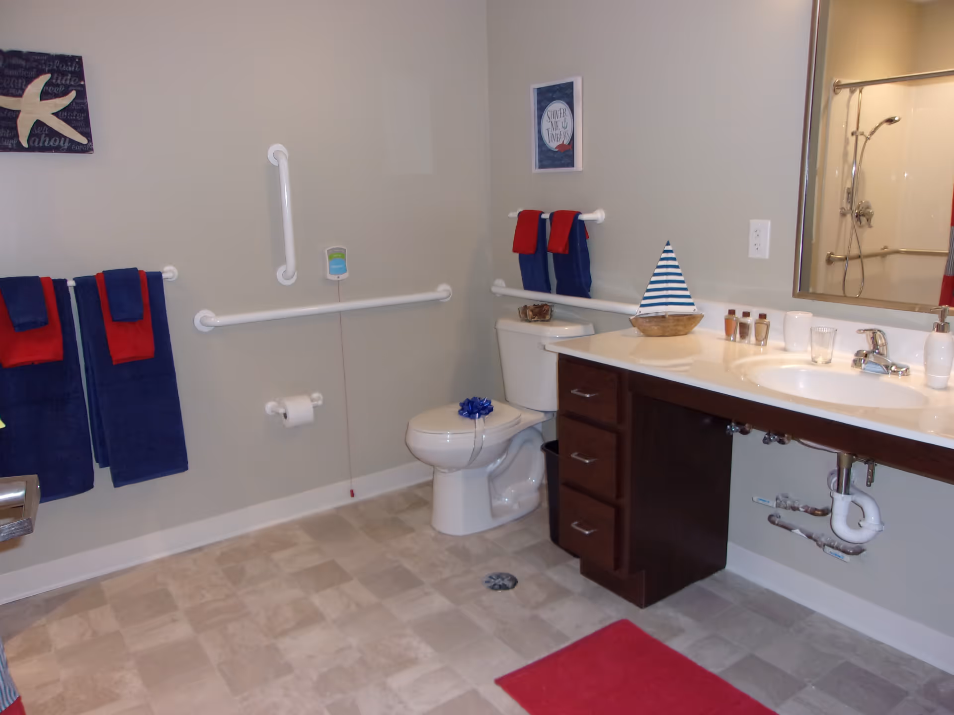 A clean and accessible bathroom with beige walls and tiled floor. The bathroom features a white toilet with grab bars on the walls around it, a white sink with a dark wood cabinet underneath, and a large mirror above the sink. There are blue and red towels hanging on towel racks, a small decorative sailboat on the counter, and a shower visible in the mirror reflection.