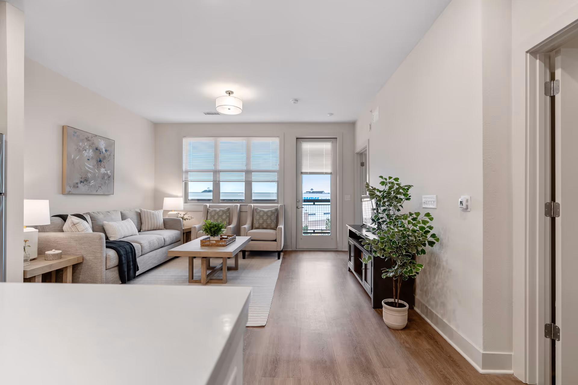 A bright and modern living room with a beige sofa, two armchairs, a wooden coffee table, and a TV stand with a potted plant. The room has large windows with blinds and a door leading outside. The floor is wooden, and the walls are painted white.
