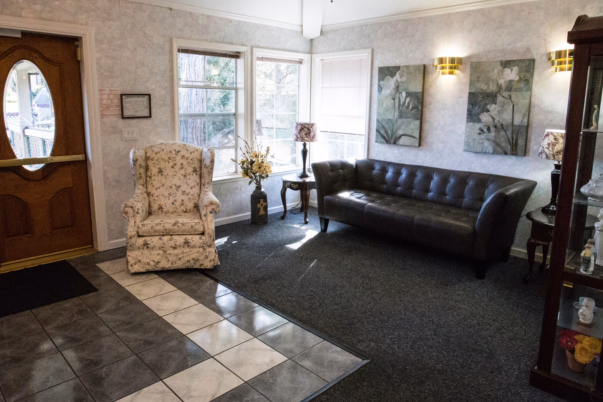 A cozy indoor lounge area with a floral armchair, a black tufted leather sofa, small side tables, lamps, and wall art by a row of windows near a wooden front door.