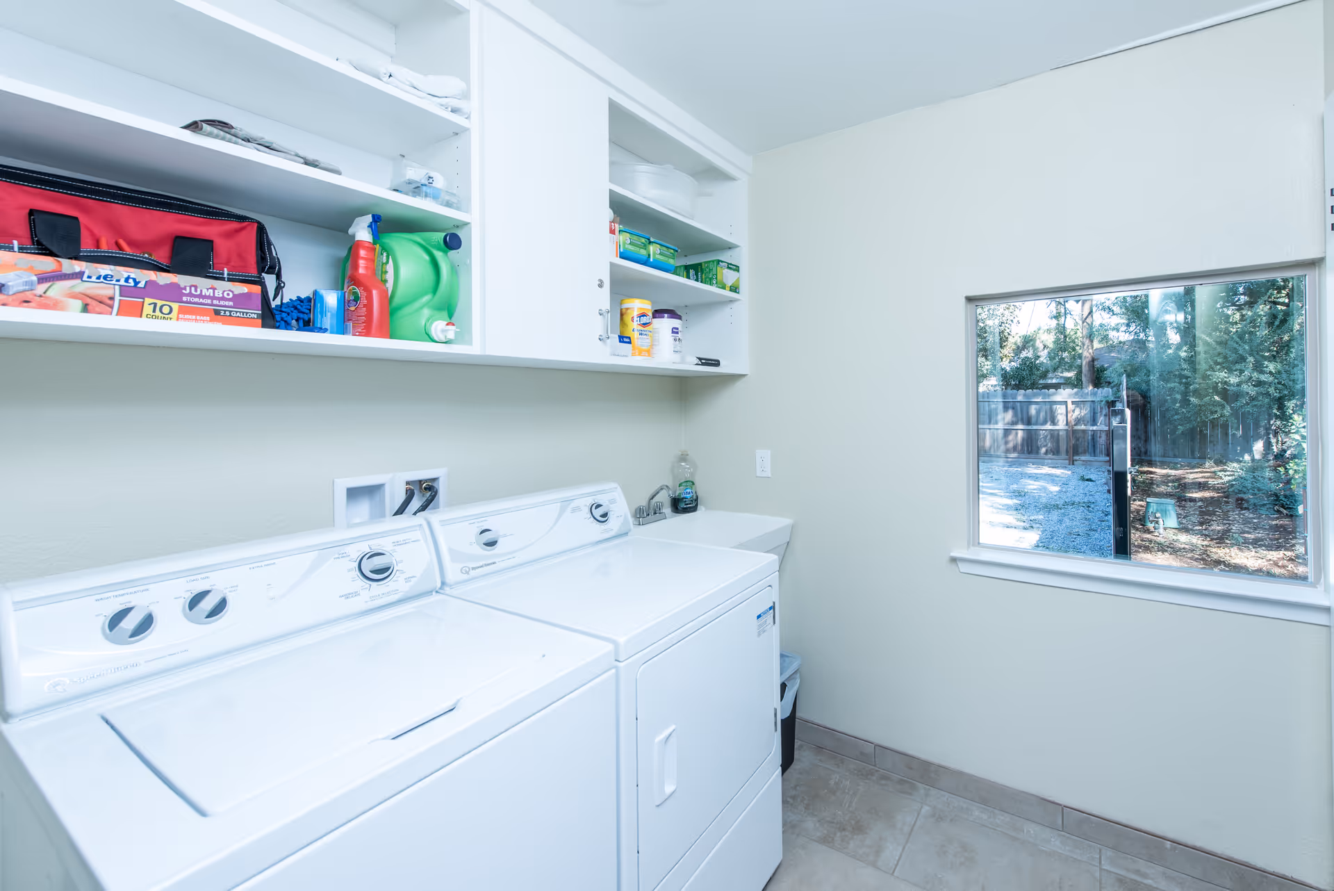Laundry room with a white washing machine and dryer side by side, white shelves above holding cleaning supplies and storage bags, a small sink in the corner, and a window showing an outdoor fenced area with trees.
