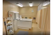 Interior view of a clean, well-lit shower room with beige tiled walls and floor. There is a white shower stall with a curtain on the right side, a white countertop with a sink and mirror in the center, and a small shelving unit on the left holding towels and toiletries.