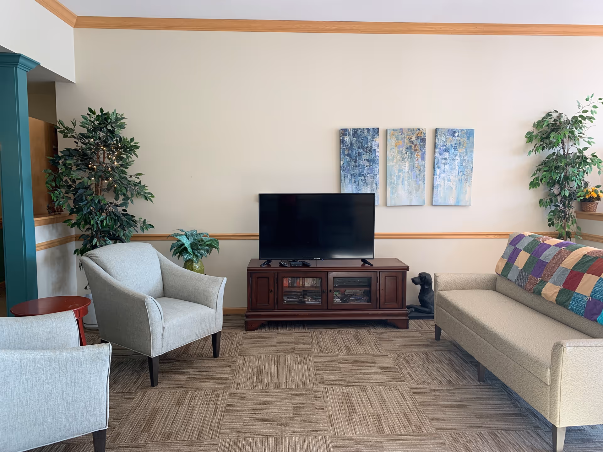 A cozy living room area with a flat-screen TV on a wooden stand, two light gray armchairs, a beige sofa with a colorful patchwork quilt draped over the back, two potted plants, and three abstract blue and white paintings on the wall.
