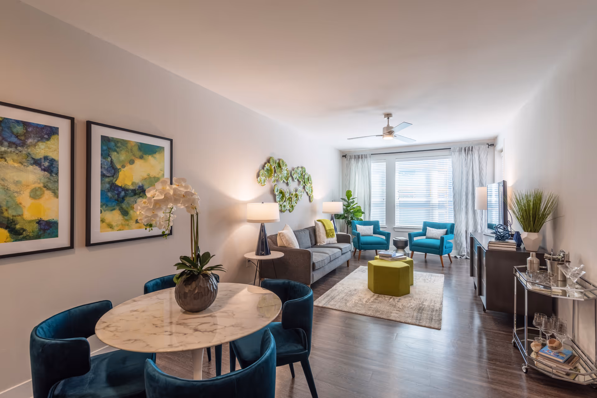 Bright modern living room and dining area with teal chairs, a gray sofa, marble round table, green ottoman, and a large window at the far end.