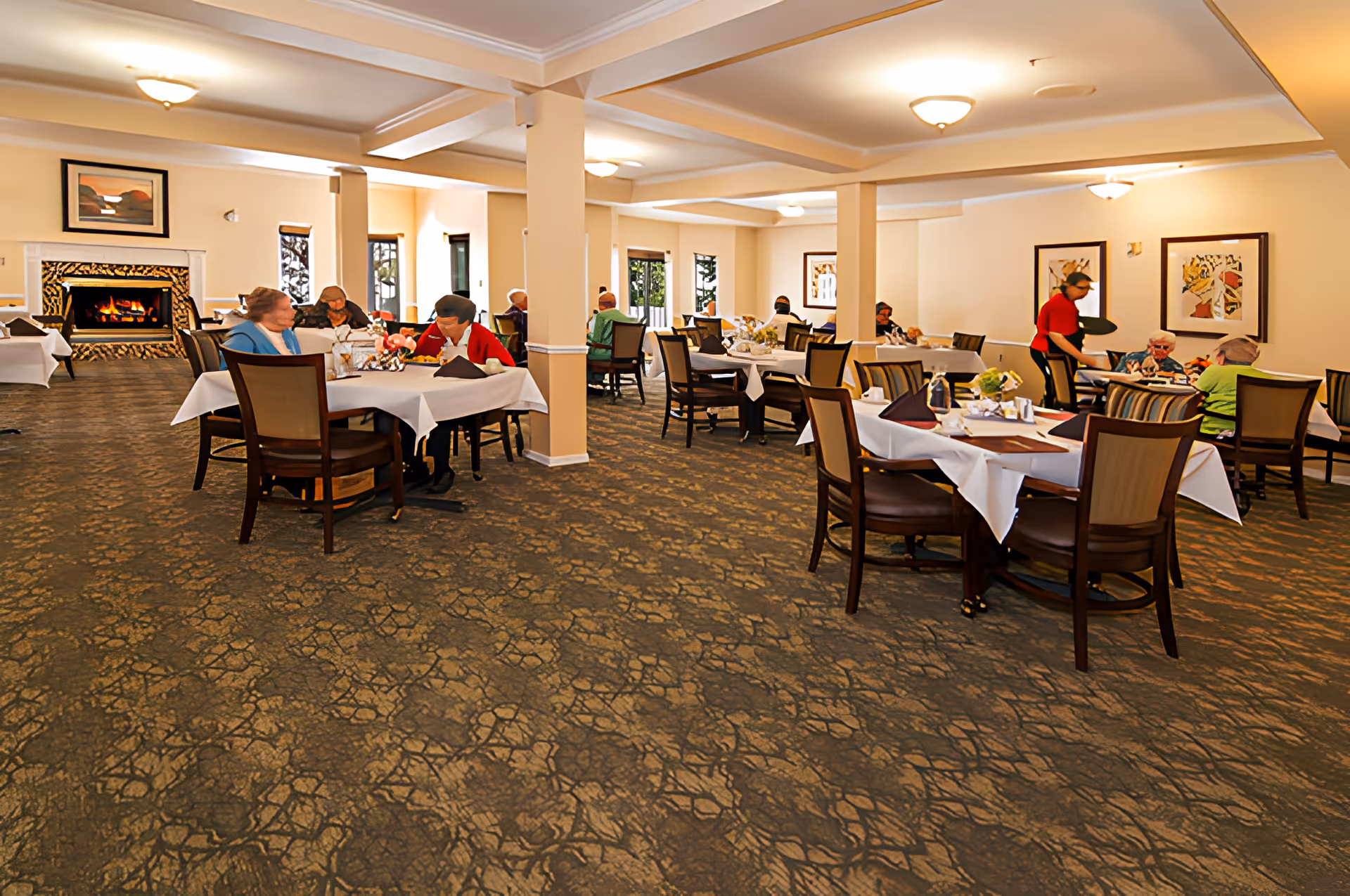 Spacious dining room with multiple tables set with white tablecloths and residents seated while staff serve them.