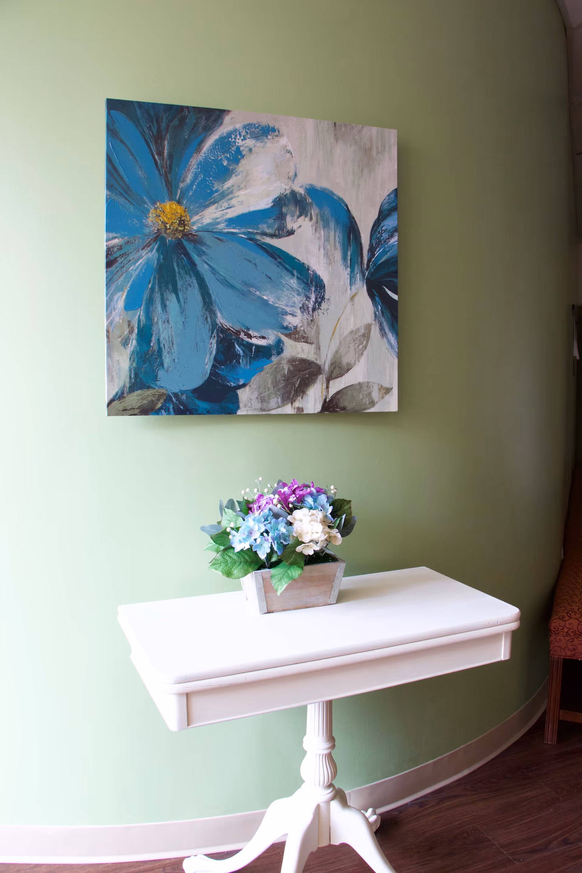 A small white wooden table with a decorative flower arrangement in a rectangular pot placed on top. Behind the table, there is a green wall with a large painting of blue and white flowers. Part of a red patterned chair is visible on the right side.