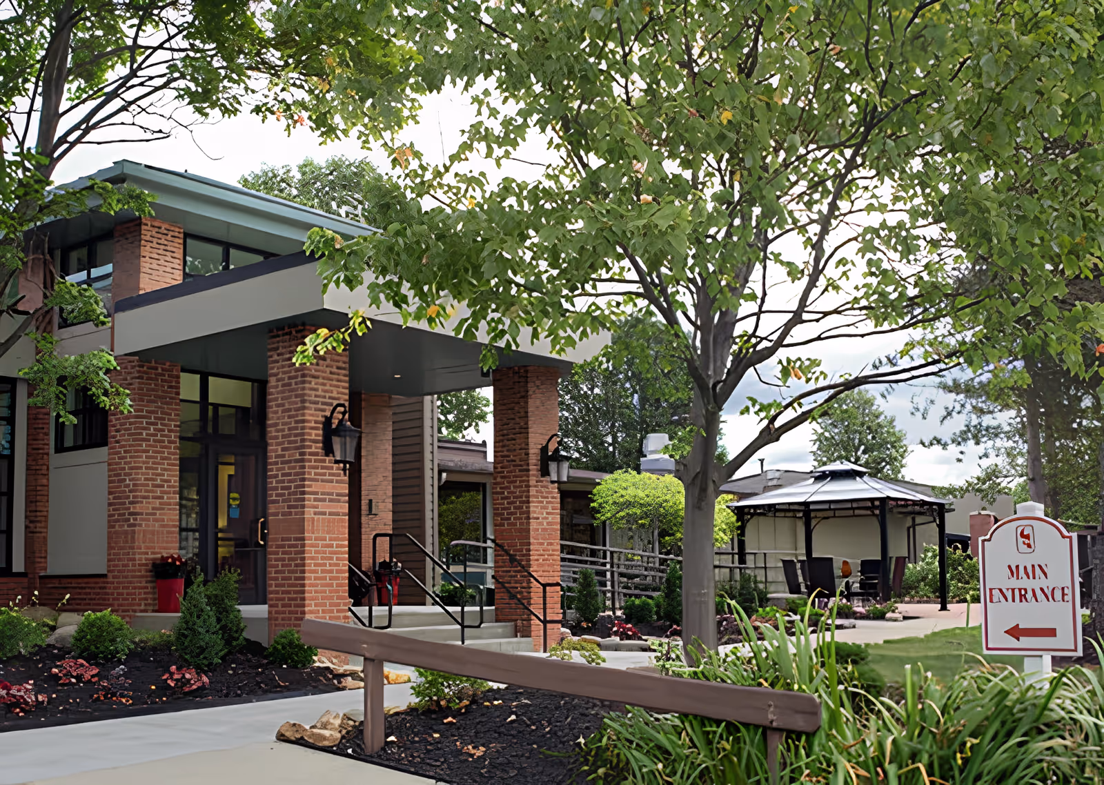 Exterior view of Cardinal Retirement Village showing the main entrance with brick pillars, a covered entryway, landscaped garden beds with shrubs and flowers, a tree in the foreground, and a gazebo with chairs in the background. A sign near the sidewalk indicates the direction to the main entrance.