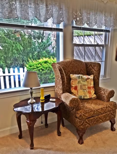A cozy corner of a living room featuring a patterned upholstered armchair with a multicolored square-patterned cushion. Next to the chair is a wooden side table holding a small lamp, a yellow flashlight, and a remote control. Behind the chair are two windows with lace valances, showing green bushes and a white picket fence outside.