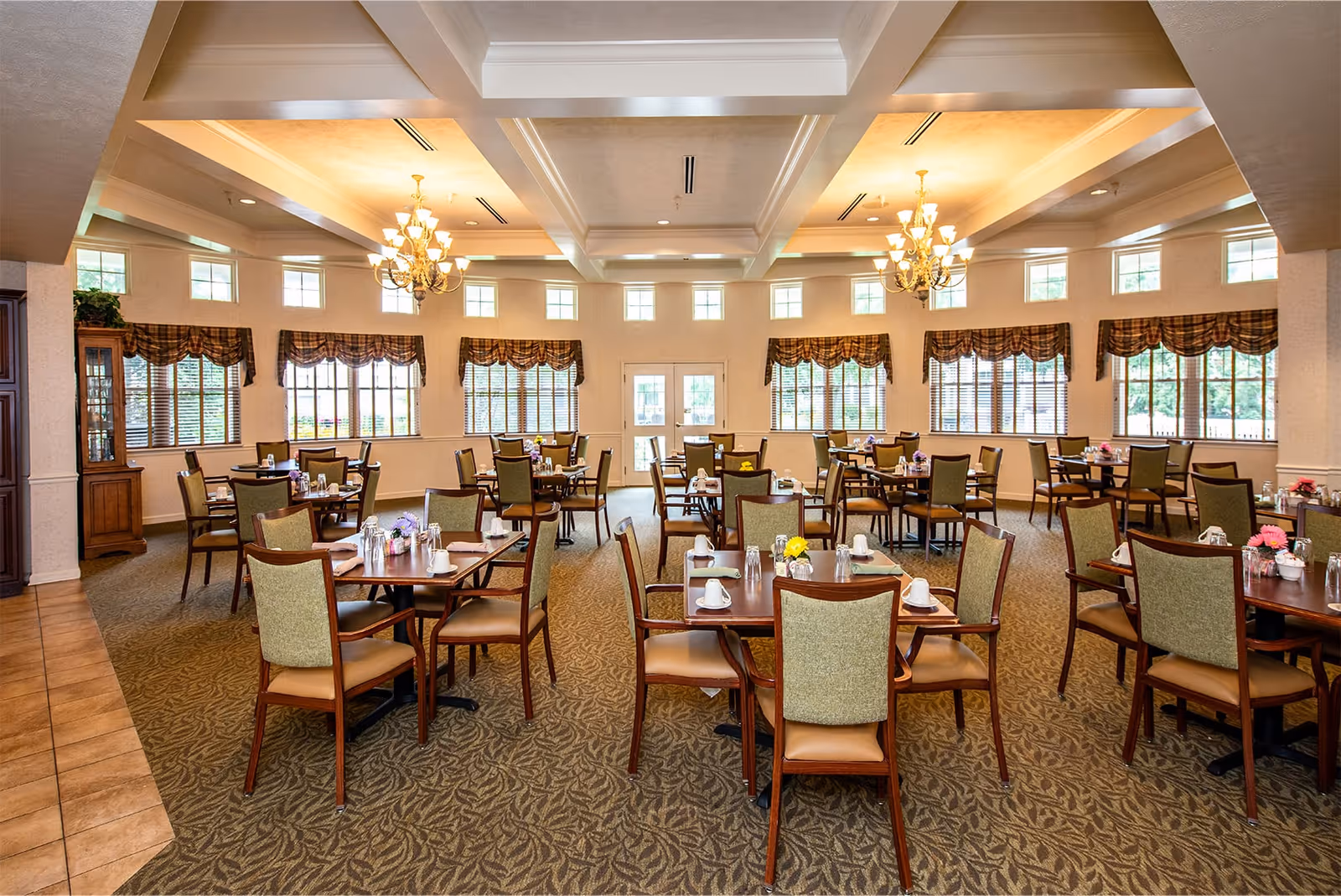 A spacious dining room with multiple wooden tables and chairs arranged neatly. Each table is set with white cups, saucers, and small flower vases. The room features large windows with patterned valances, allowing natural light to fill the space. Two elegant chandeliers hang from the coffered ceiling, and the floor is carpeted with a patterned design.
