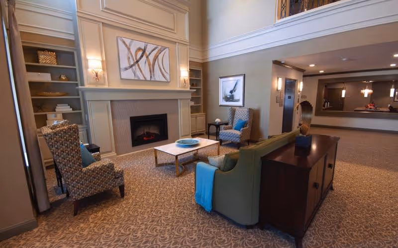 A cozy living room area in a senior living facility featuring a fireplace with abstract art above it, two patterned armchairs with blue cushions, a green sofa with a blue throw, a coffee table with a decorative bowl, built-in shelves with decorative items, and a console table with a sculpture. The room has carpeted flooring and soft lighting.