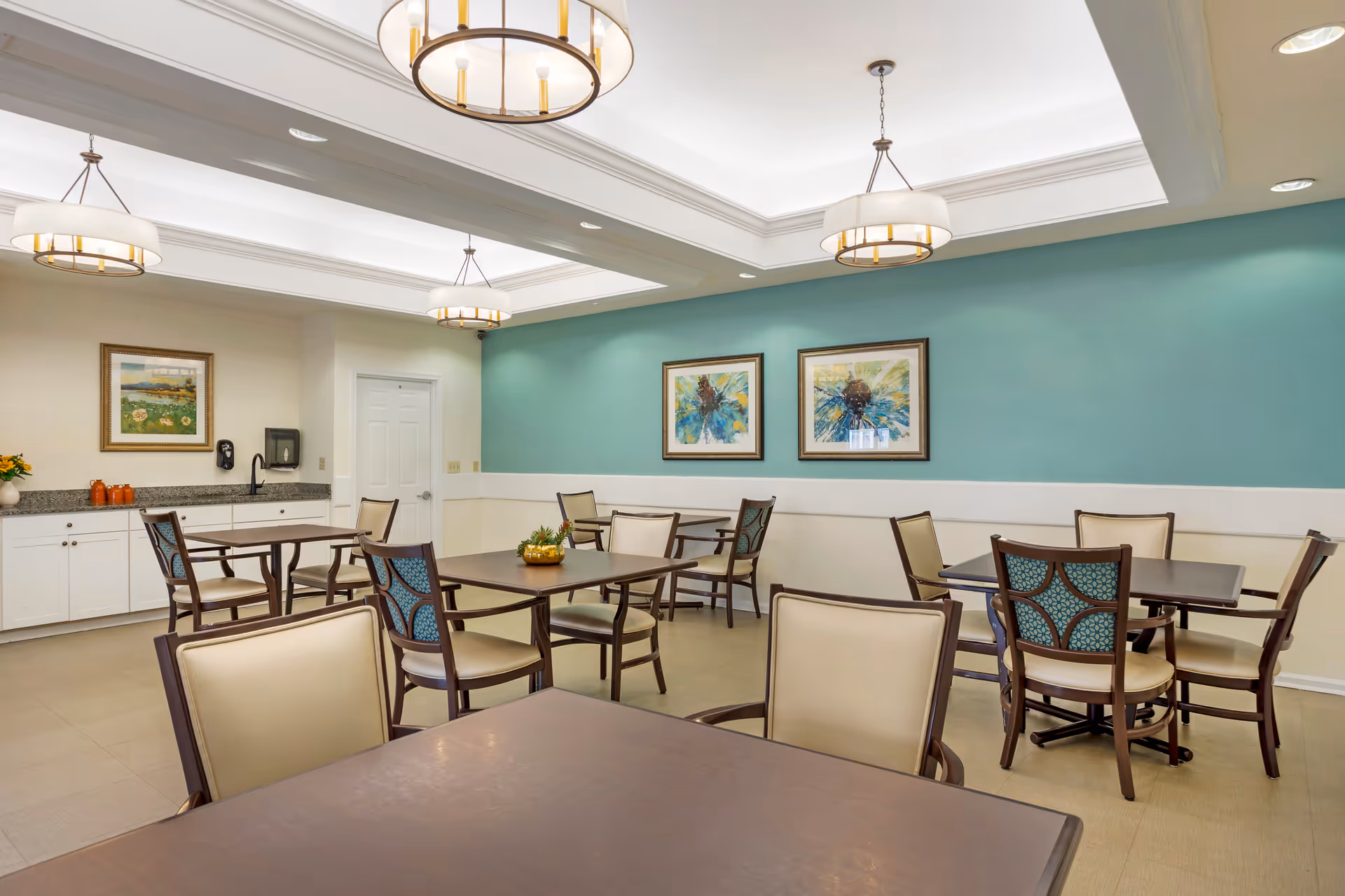 A bright and clean dining room with several tables and chairs arranged neatly. The walls are painted teal and white, adorned with two framed abstract paintings. There are three modern pendant lights hanging from the ceiling, and a countertop with cabinets and a sink is visible in the background.