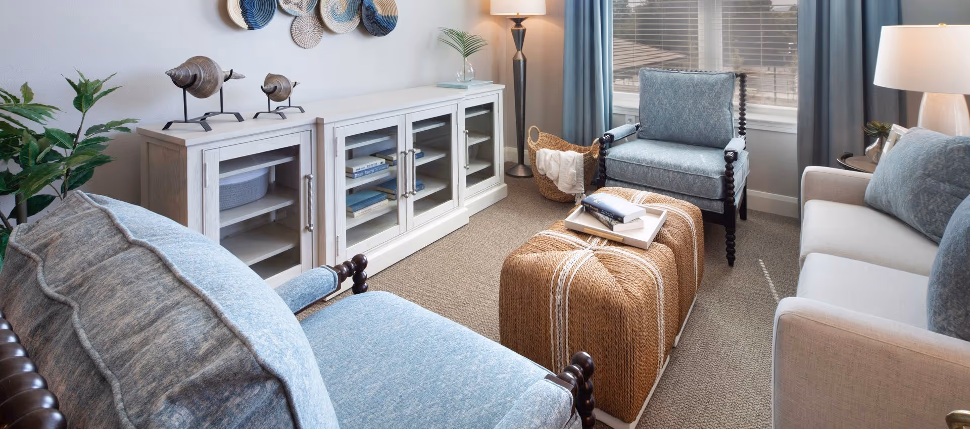 A cozy living room with two blue upholstered armchairs, a beige sofa with blue cushions, a woven ottoman serving as a coffee table with books on a tray, a white cabinet with glass doors containing books, decorative wall hangings, a floor lamp, and a window with blue curtains.