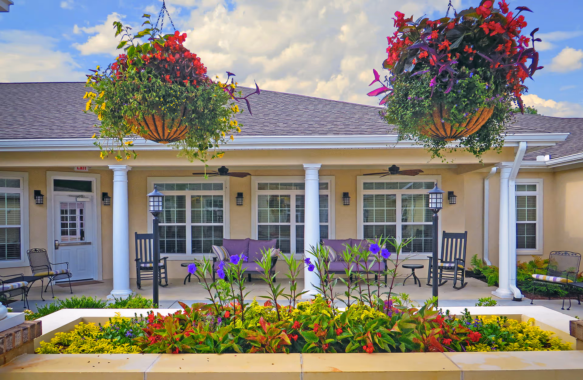 Outdoor patio area of Benton Manor Of Stockbridge featuring a flower bed with colorful flowers in the foreground, two hanging flower baskets with red and purple flowers, white columns supporting the roof, rocking chairs, and benches with cushions on the porch under a partly cloudy sky.