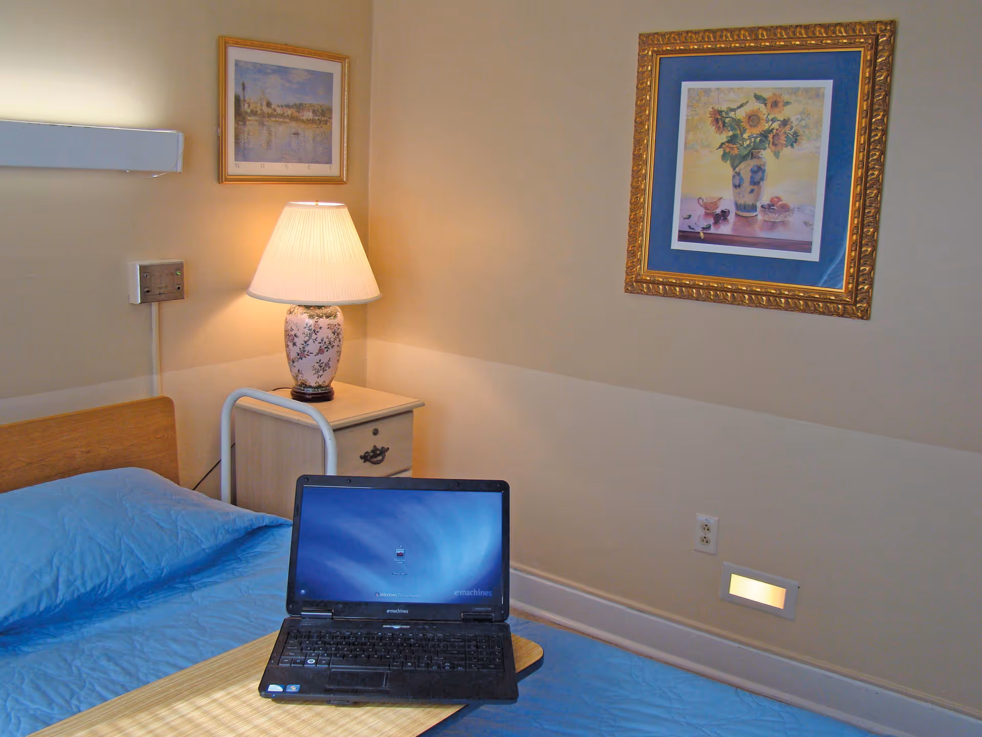A nursing center bedroom with a single bed covered in a blue bedspread. On the bed is a laptop computer. Next to the bed is a nightstand with a floral table lamp. The walls are beige and decorated with two framed pictures, one of a landscape and the other of a vase with sunflowers.