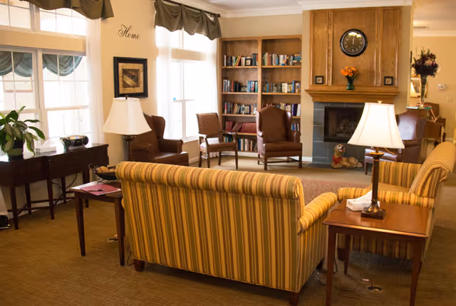 A cozy living room area with a striped yellow sofa, two brown armchairs, a wooden bookshelf filled with books, a fireplace with a clock above it, and two table lamps on wooden side tables. Large windows with curtains allow natural light to fill the room, and there is a small table with a plant and a radio near the window.