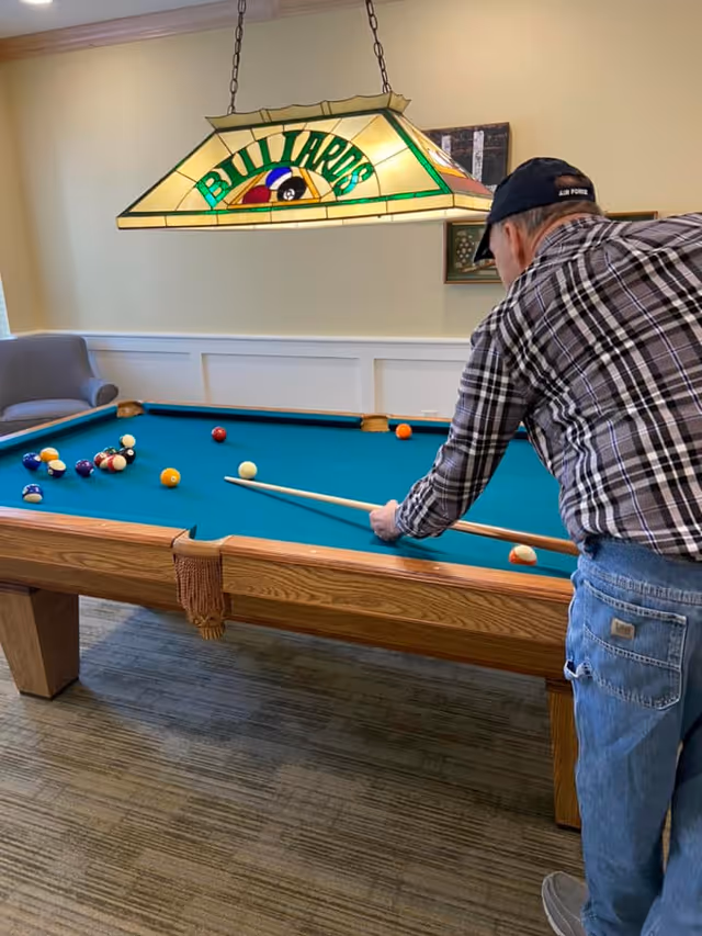 An elderly man wearing a plaid shirt and jeans is playing billiards on a pool table in a room with beige walls and carpeted floor. A stained glass light fixture above the table displays the word 'BILLIARDS'. There is a gray armchair in the corner and framed artwork on the wall.