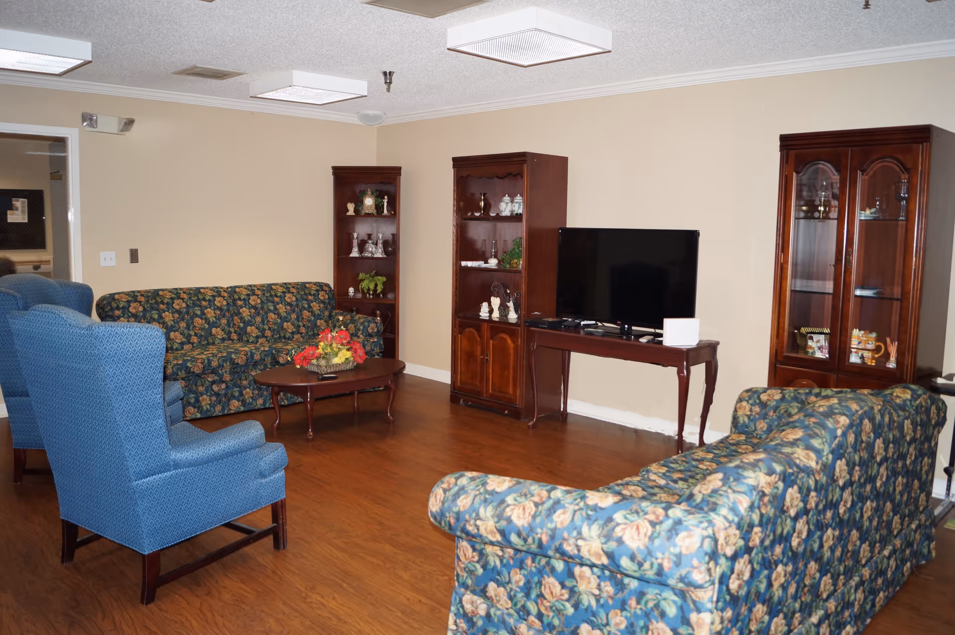 A living room area with floral patterned sofas and blue armchairs arranged around a wooden coffee table with a flower arrangement. There are wooden display cabinets and a TV on a wooden table against a beige wall. The floor is wooden and the ceiling has rectangular fluorescent lights.
