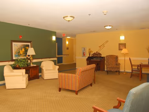 A spacious senior living facility common area with beige carpet and walls painted green and yellow. The room contains several upholstered chairs and a plaid sofa arranged around a wooden side table with a lamp and a plant. There is a framed painting on the green wall and a decorative trombone mounted on the yellow wall above a small wooden cabinet. Additional seating and a table are visible in the background.