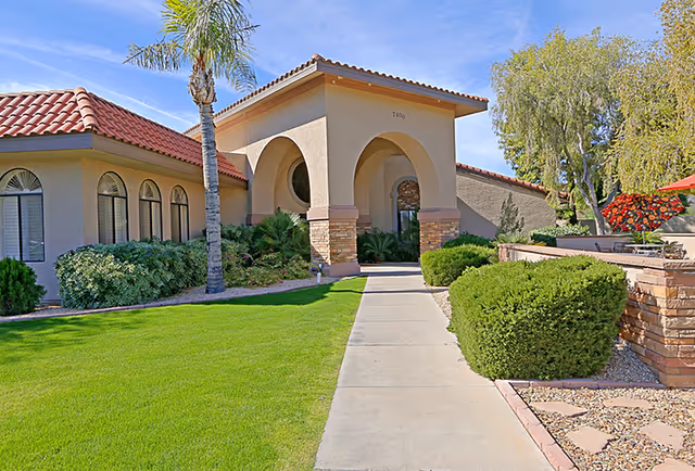 Exterior view of a senior living facility with a beige stucco building featuring a red tile roof, arched entryway, and well-maintained landscaping including green grass, bushes, palm trees, and a stone wall with red flowers and an umbrella in the background.
