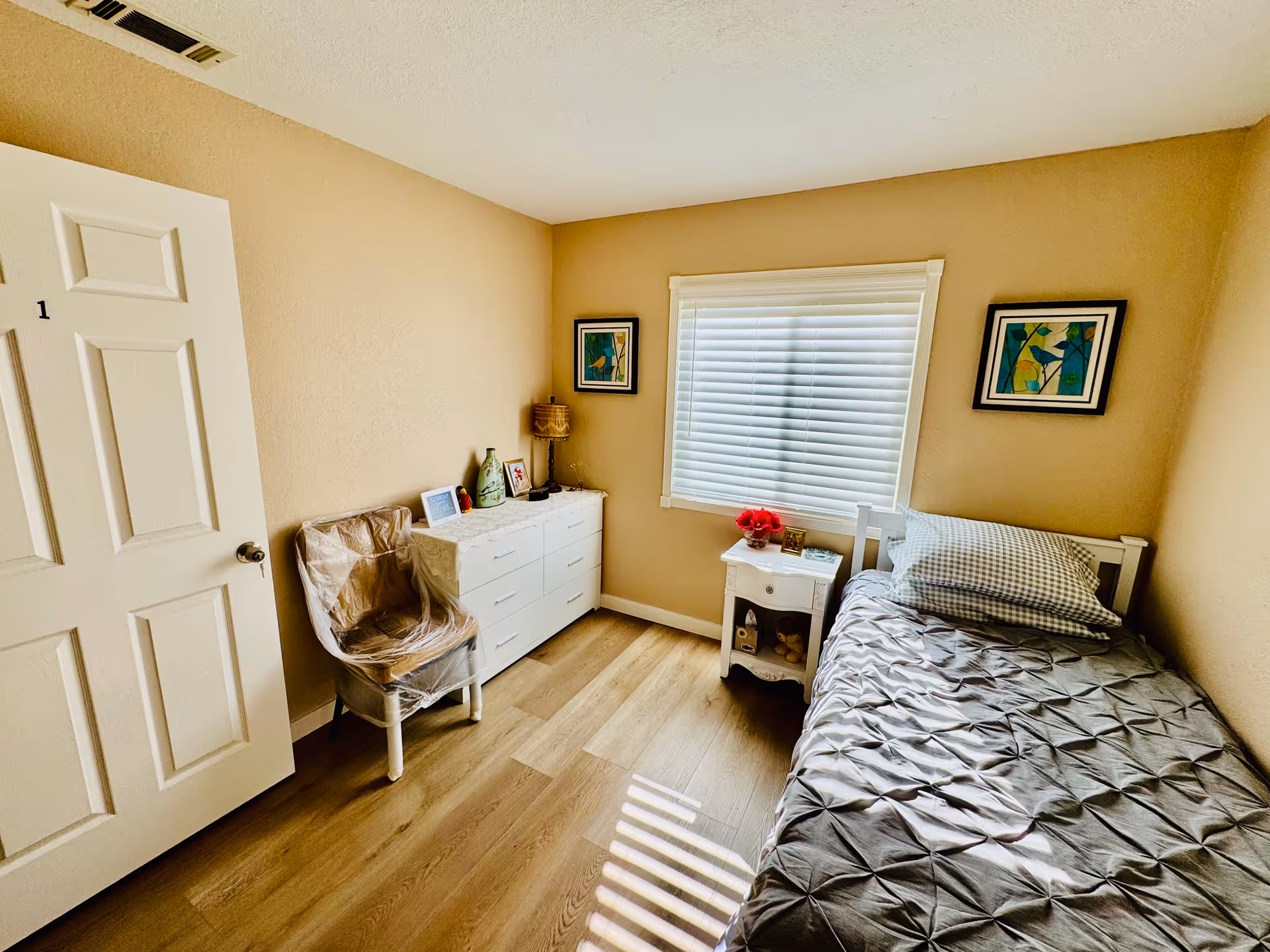 A small, cozy bedroom with a single bed covered in a gray comforter and checkered pillows. There is a white nightstand next to the bed with a small flower vase and picture frame on top. A white dresser with several drawers is against the opposite wall, with a chair covered in plastic beside it. Two framed pictures hang on the beige walls, and a window with white blinds lets in natural light.