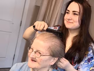 A young woman is brushing the hair of an elderly woman who is seated and wearing glasses, inside a room with a closed door and curtains in the background.