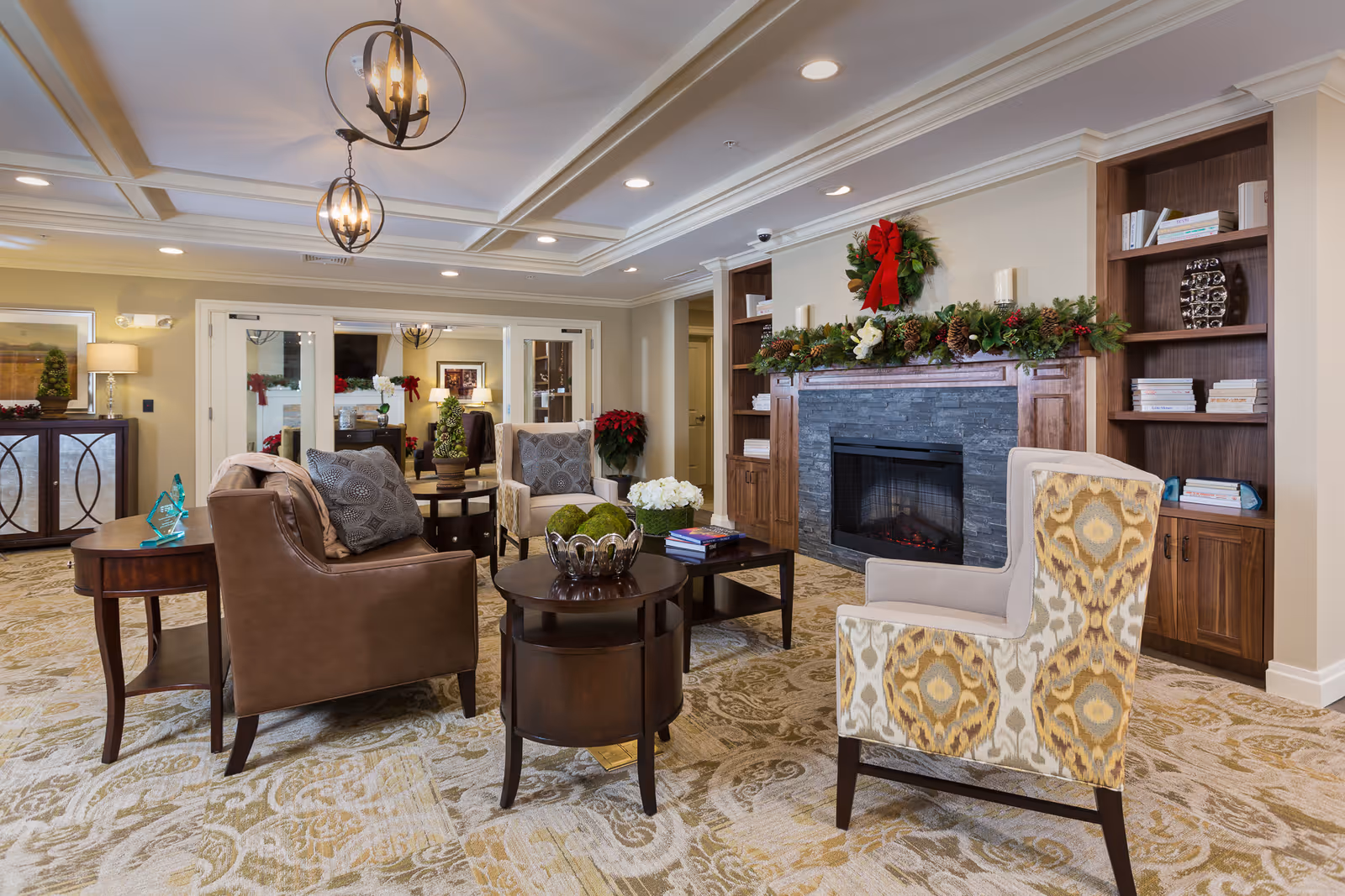 A cozy living room area with a fireplace decorated with a Christmas wreath and garland. The room features comfortable seating including a brown leather sofa, patterned armchairs, and wooden side tables. Built-in wooden bookshelves flank the fireplace, and the ceiling has recessed lighting and decorative light fixtures. The carpet has an intricate pattern, and there are holiday decorations such as poinsettias and a small Christmas tree in the background.