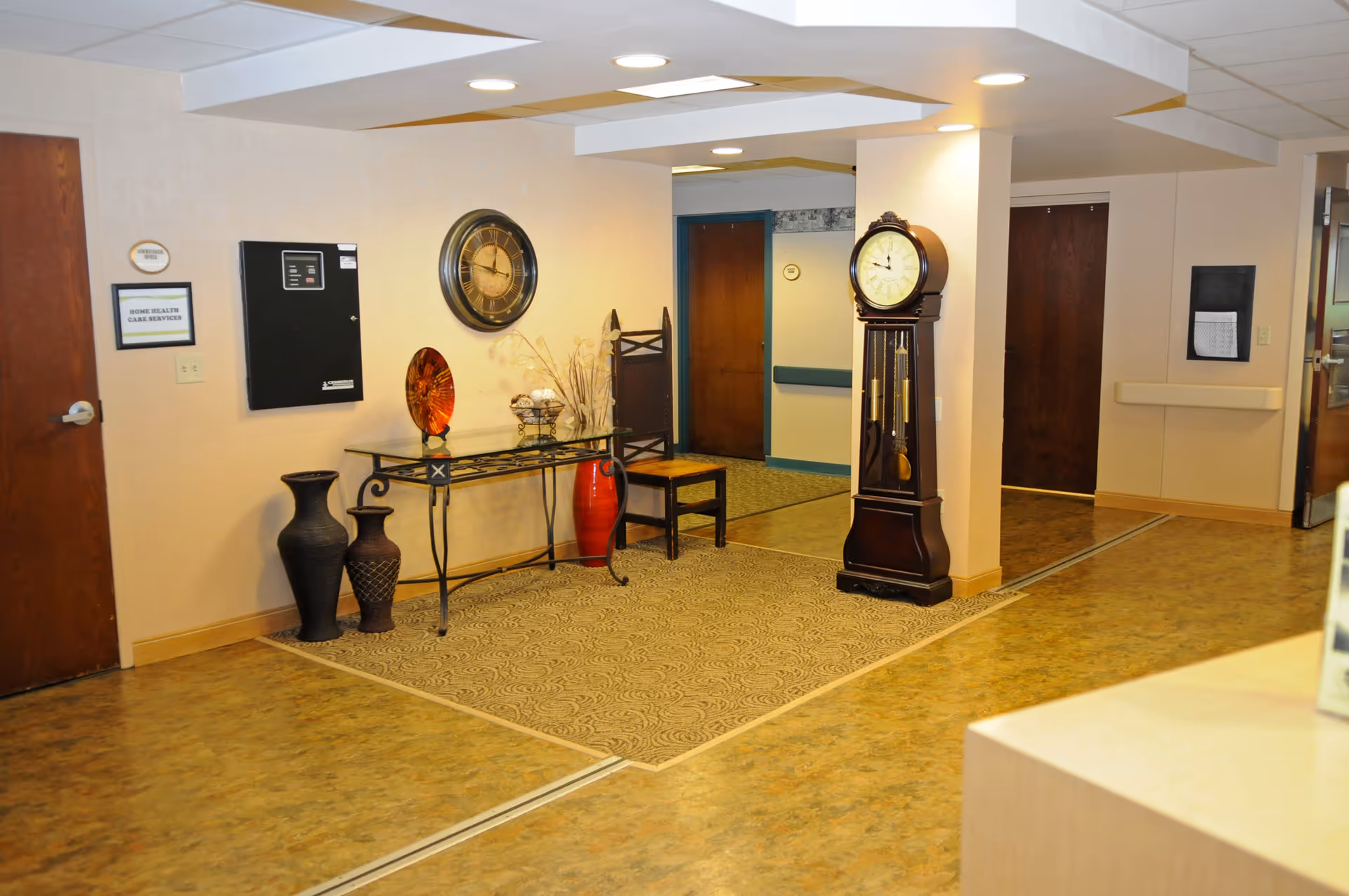 Interior hallway area of a senior living facility with a decorative glass table holding a red vase, a bowl with decorative balls, and dried plants. There are two large vases on the floor next to the table, a wooden chair, a large wall clock, and a tall grandfather clock. Several wooden doors and a beige carpet with a patterned design are visible.