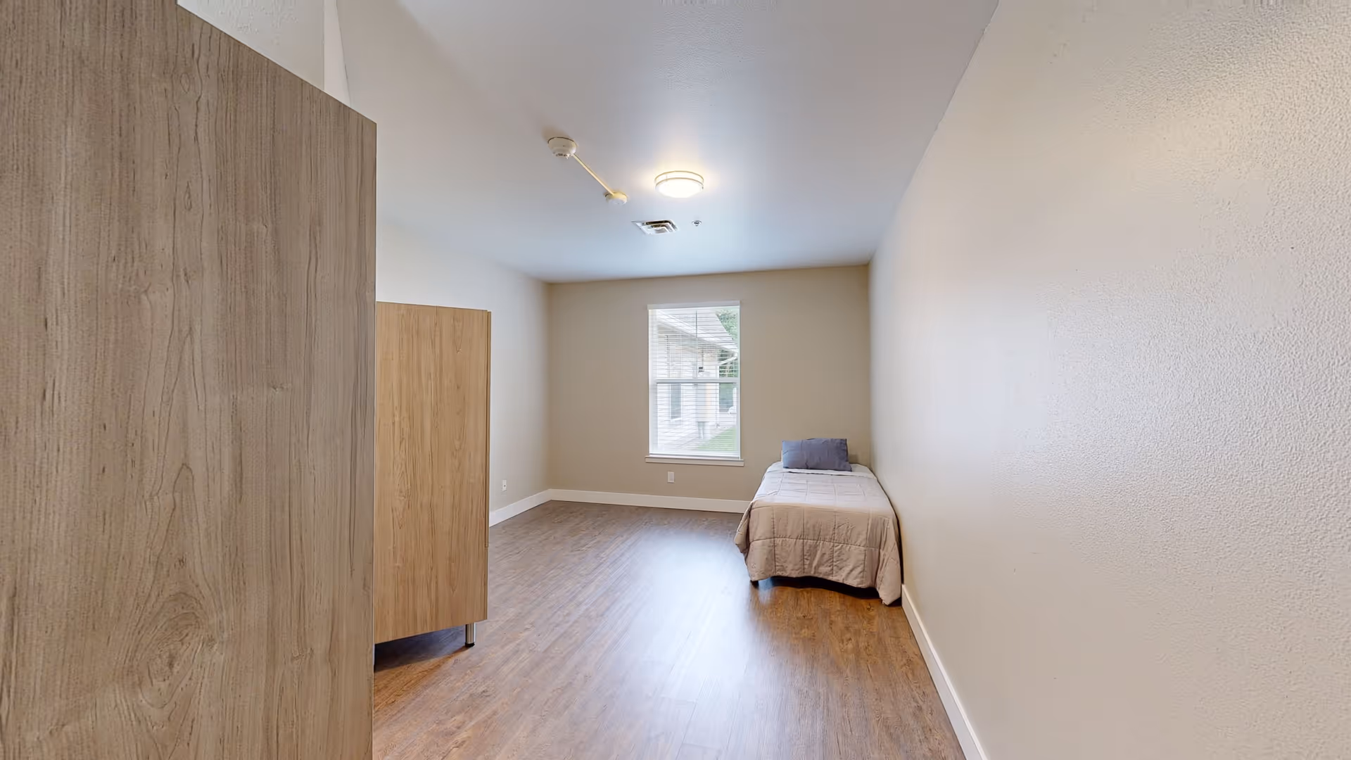 Simple bedroom with a single bed, wooden wardrobe, a window, and wood flooring.