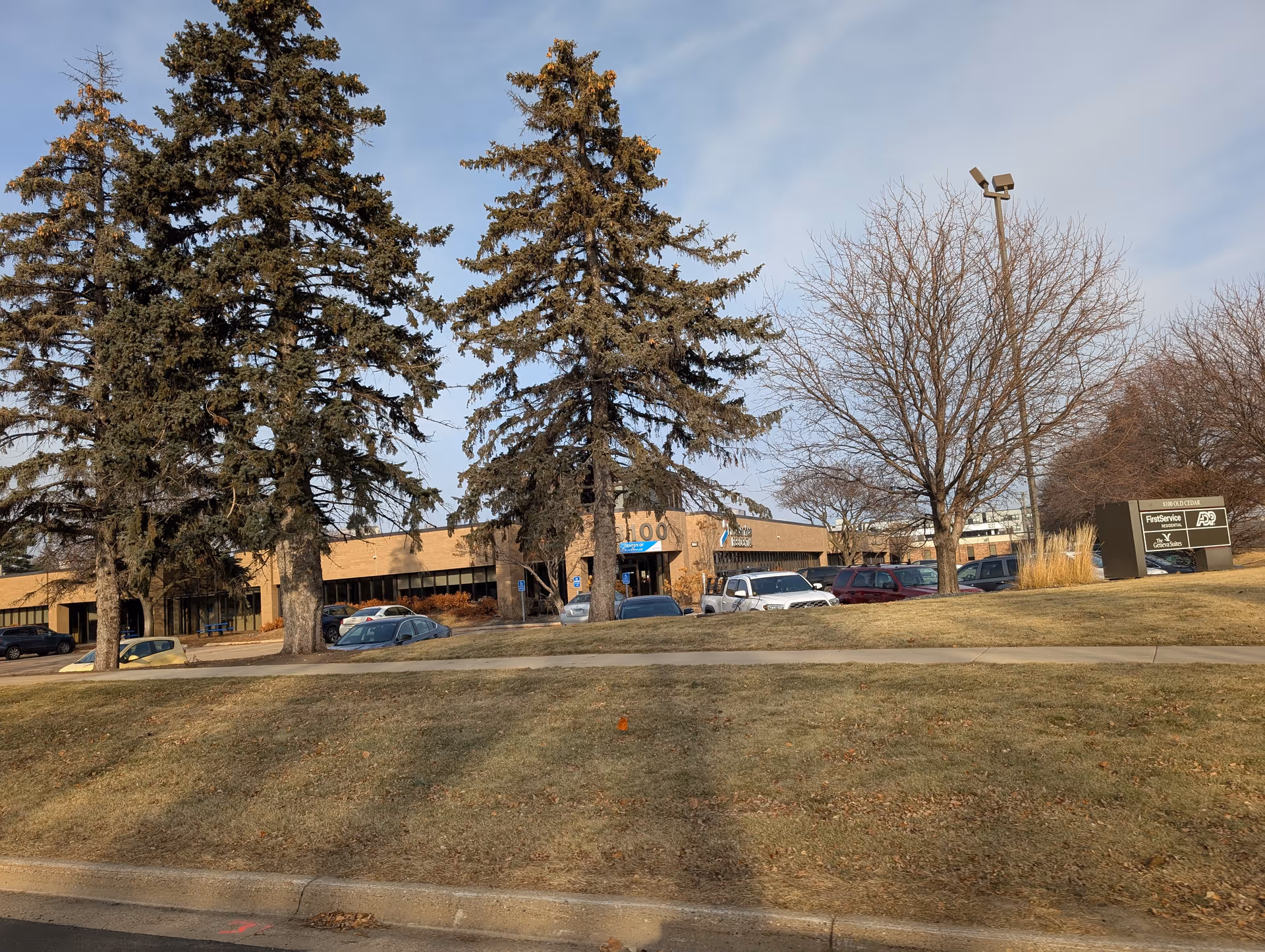 Exterior view of a single-story brick building with several tall evergreen trees and leafless deciduous trees in front. There is a parking lot with multiple cars parked and a sign on the right side displaying 'The Geneva Suites' along with other business names. The sky is clear with some light clouds.