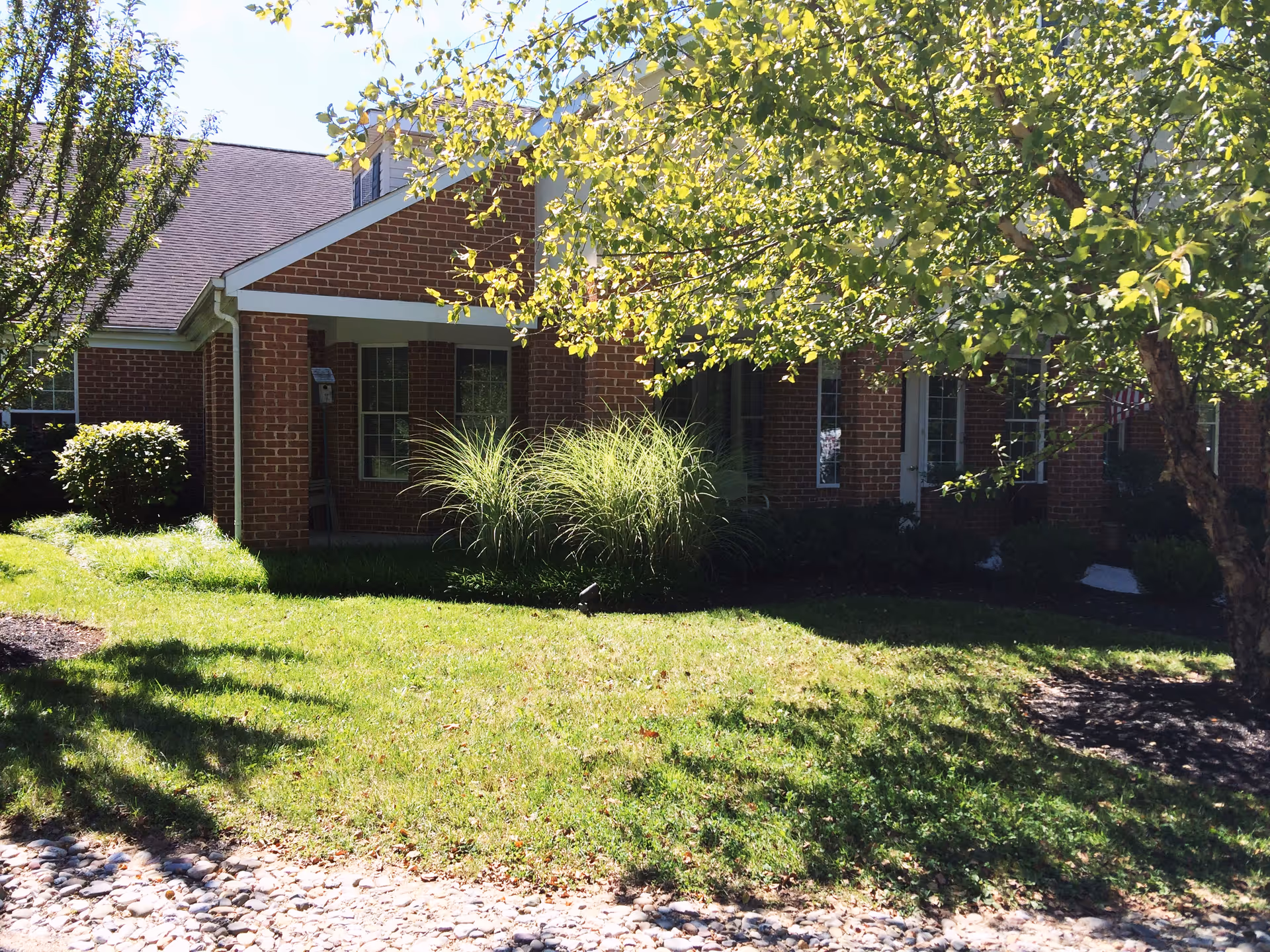 Exterior view of a brick building with a covered porch, surrounded by green grass, bushes, and trees with sunlight filtering through the leaves.