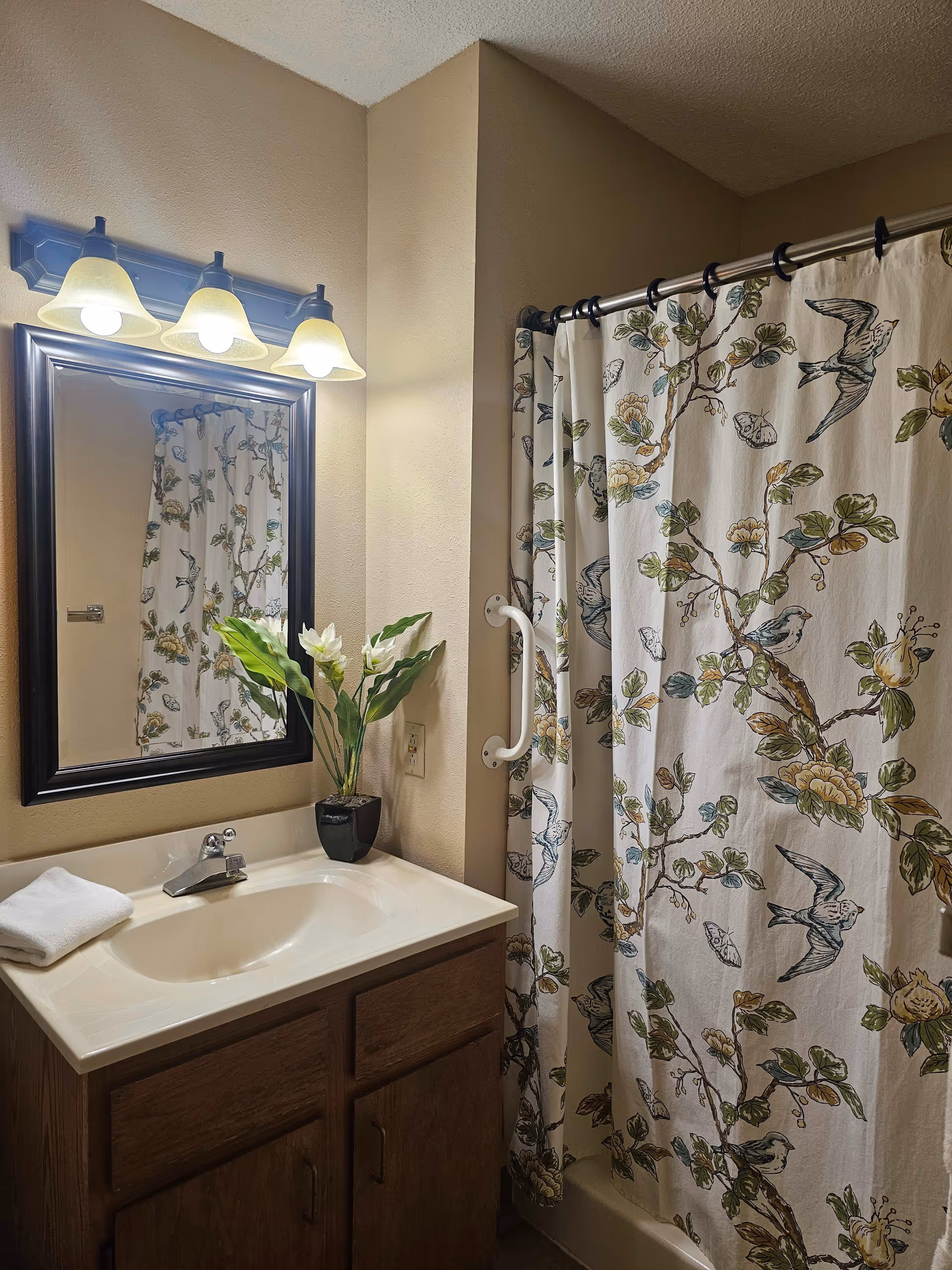 A bathroom with a sink and wooden cabinet below, a mirror with a black frame above the sink, and a three-light fixture with frosted glass shades. A white towel is folded on the sink countertop next to a small black vase with green leaves and white flowers. To the right, there is a shower with a curtain featuring a floral and bird pattern, and a white grab bar is mounted on the wall next to the shower.