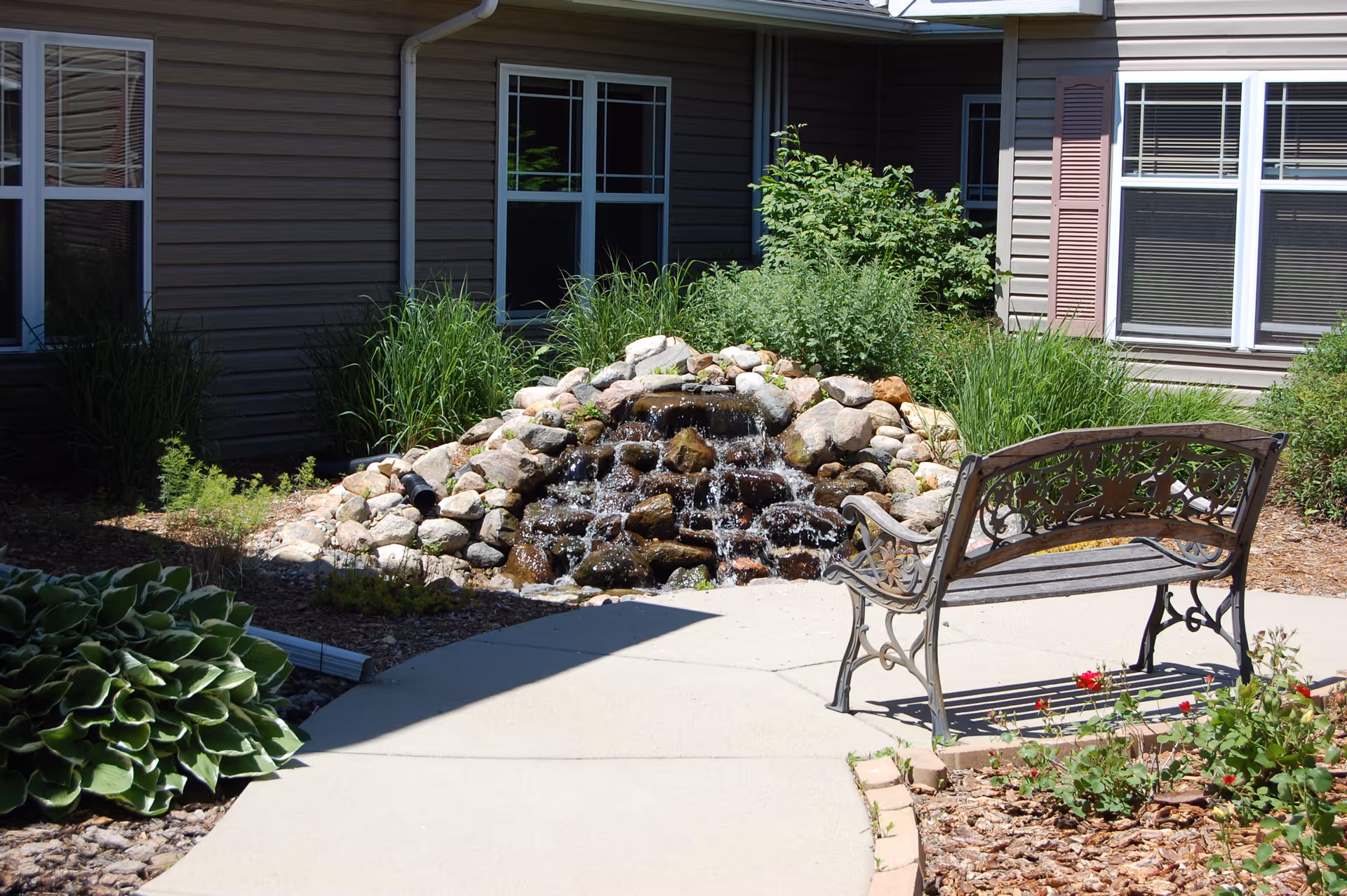 Outdoor garden area at StoneyBrook Suites Assisted Living featuring a small rock waterfall surrounded by green plants and bushes, a curved concrete pathway, and a decorative metal bench facing the waterfall. The background shows the exterior walls and windows of the facility building.