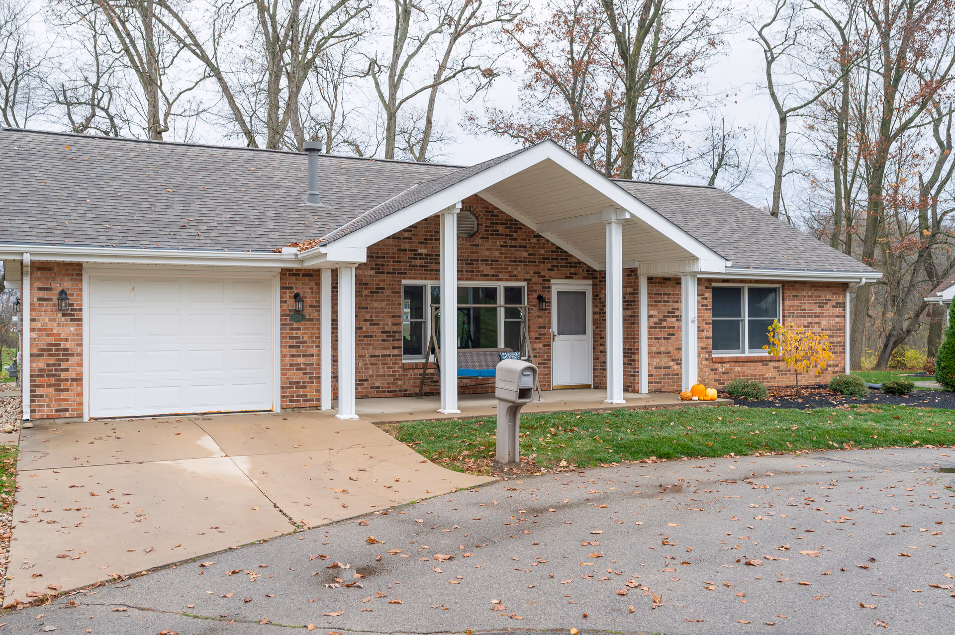 Single-story brick building with a white garage door, a covered front porch supported by white columns, a white front door, and windows. There is a mailbox on the lawn, some pumpkins near the porch, and leafless trees in the background indicating late autumn.