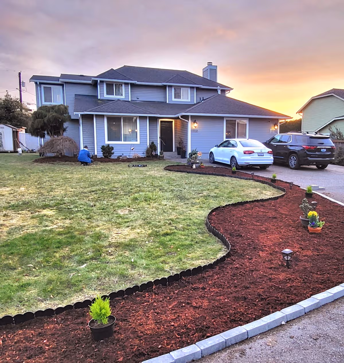 A two-story gray house with a well-maintained front yard at sunset. The yard features a curved flower bed with red mulch and small potted plants along the edge. Two cars are parked in the driveway, and a person wearing a blue jacket is working in the garden near the house.