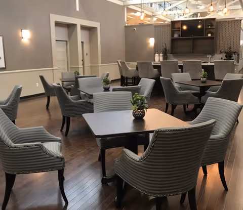 Spacious dining/lounge area with multiple dark wood tables and upholstered chairs on hardwood floors, small potted plants on the tables, and a service counter in the background.