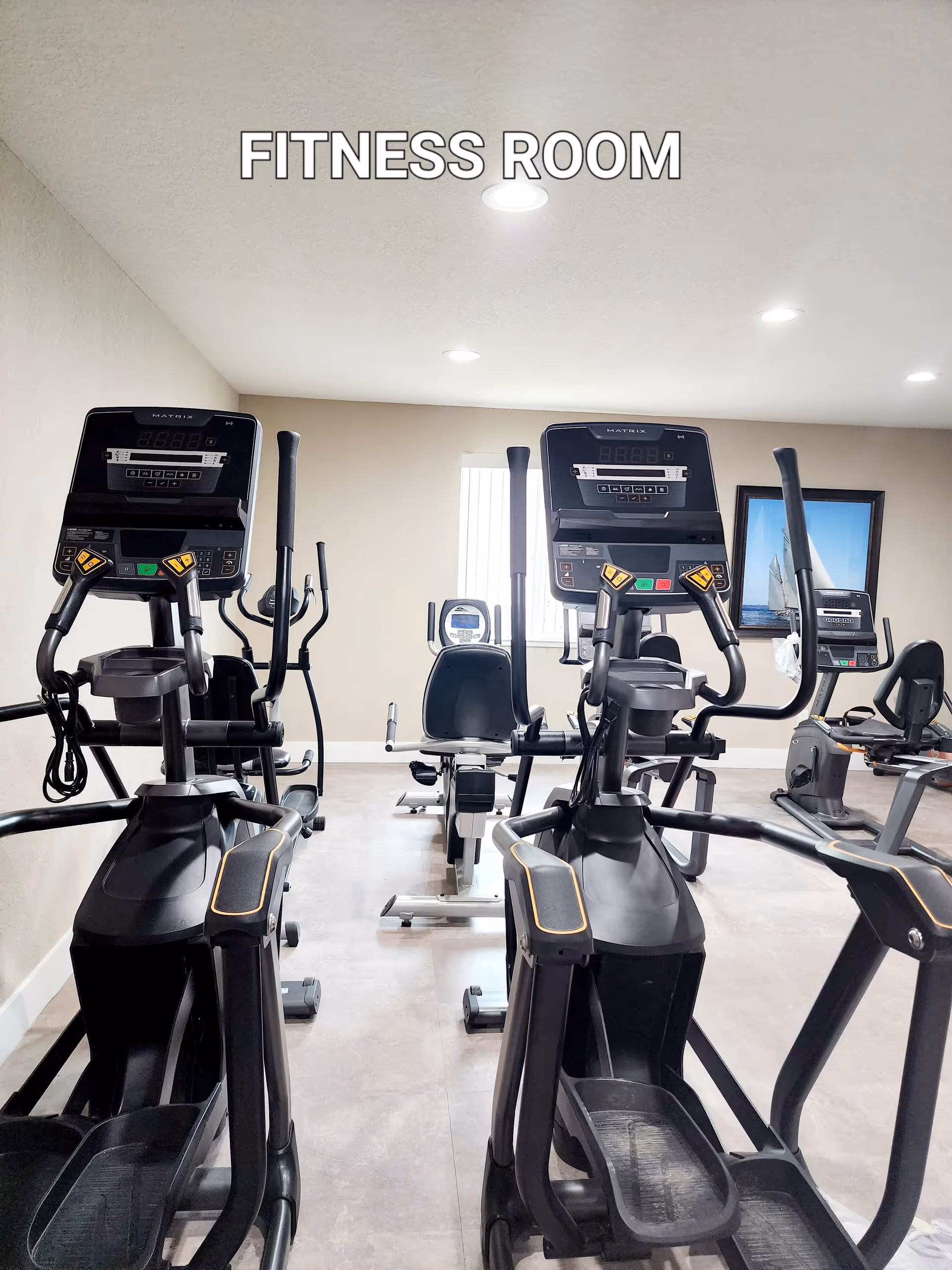 Fitness room with several exercise machines including elliptical trainers and stationary bikes, beige walls, a window, and a framed picture of a sailboat on the wall.