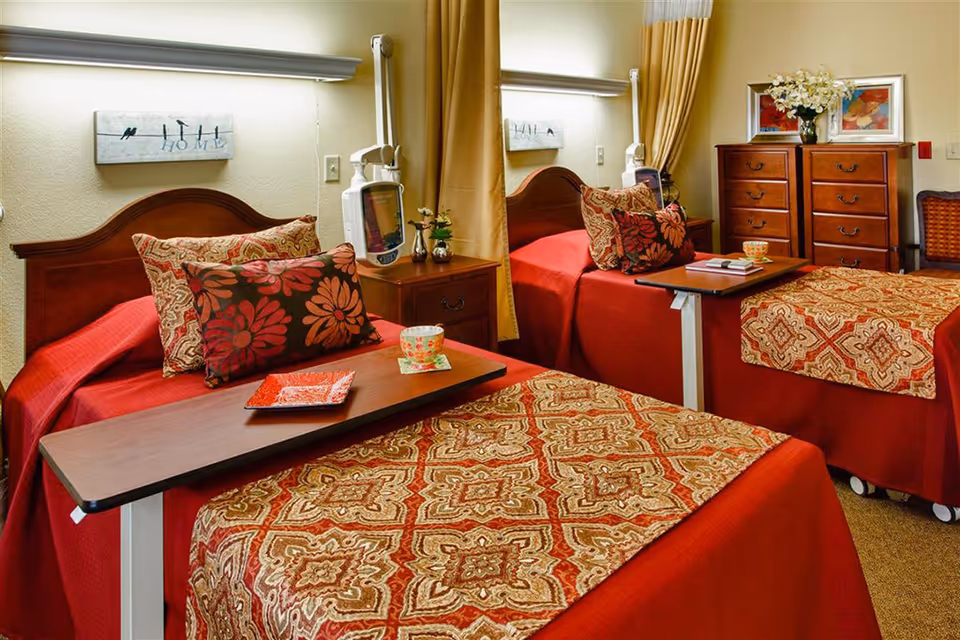 A cozy bedroom in a senior living facility featuring two single beds with red and patterned bedspreads and decorative pillows. Each bed has an overbed table with a cup and plate. Between the beds is a nightstand with a small plant and a medical device mounted on the wall. A wooden dresser with flowers and framed artwork is visible in the background. The room has warm lighting and beige walls with curtains.