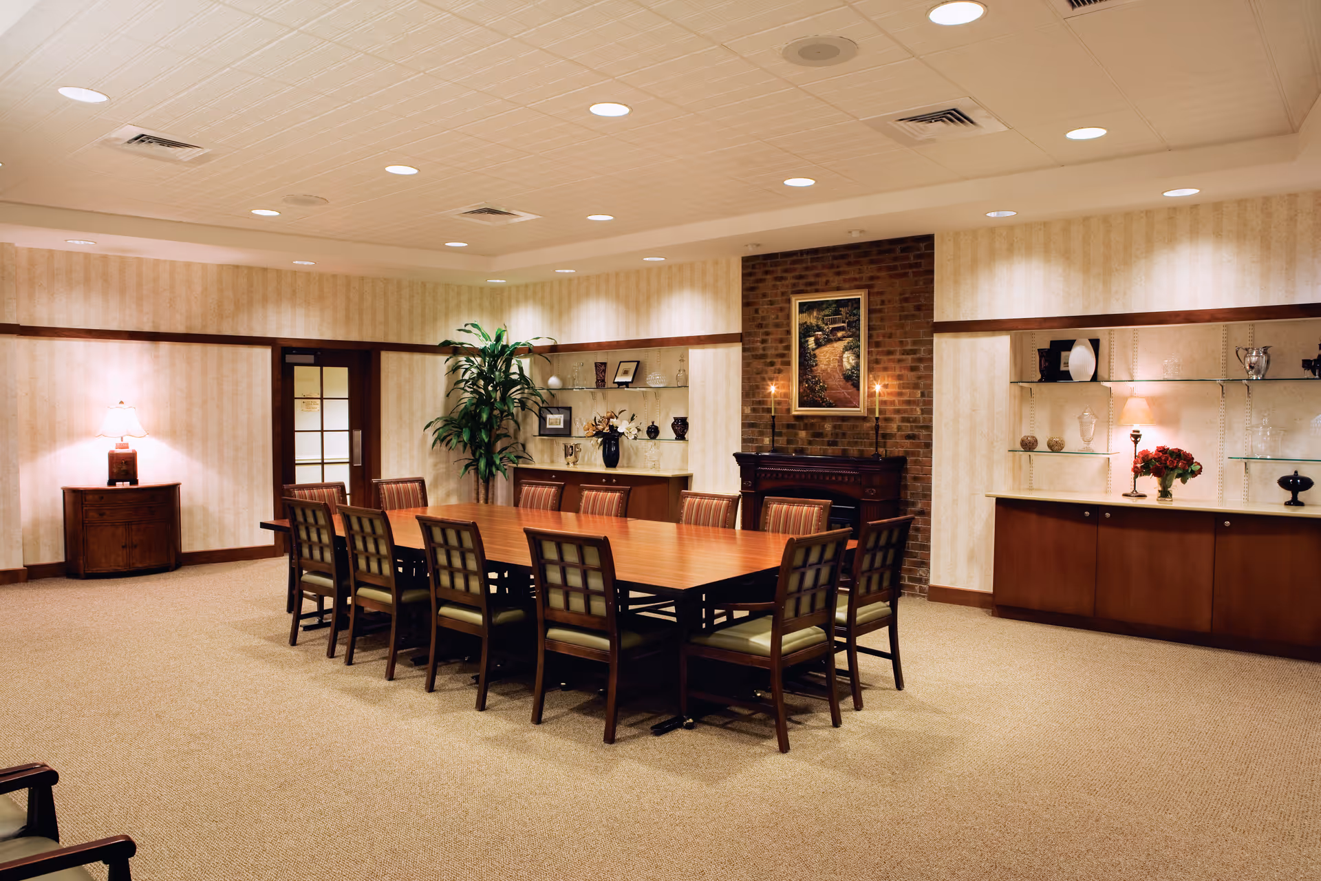 A well-lit meeting or dining room with a long wooden table surrounded by ten chairs. The room features beige carpet, wallpapered walls with a wooden trim, a brick fireplace with two lit candles and a framed picture above it, built-in shelves with decorative items, a potted plant, and a small wooden cabinet with a lamp on it.