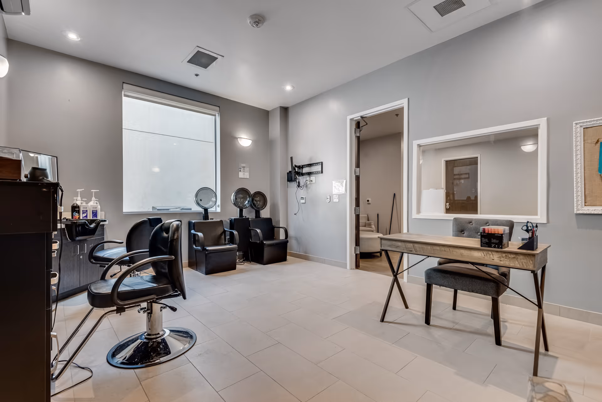 Interior view of a salon area in a senior living facility with black salon chairs, hair drying stations, a wooden desk with a gray chair, and a large window letting in natural light.