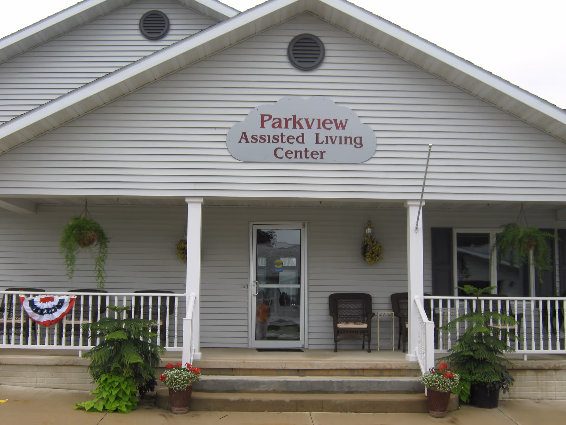Front exterior view of Parkview Assisted Living Center with a porch featuring two wicker chairs, hanging plants, potted flowers, and a patriotic bunting decoration on the railing.