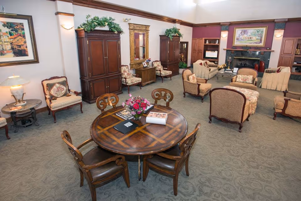 A spacious living room area with multiple upholstered armchairs and sofas arranged around a fireplace. There is a round wooden table with four chairs in the foreground, decorated with a flower arrangement and some books. The room features carpeted flooring, framed artwork on the walls, and wooden cabinets with plants on top.