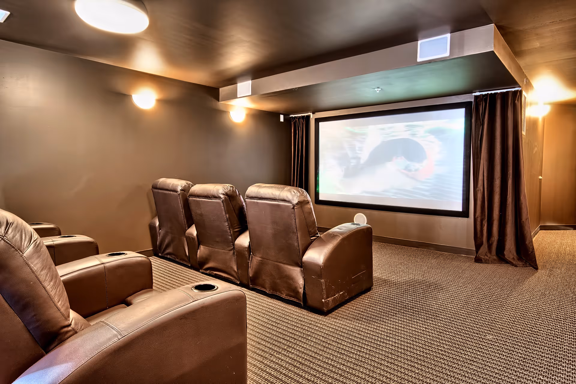 A small home theater room with six brown leather recliner chairs arranged in two rows facing a large projection screen. The room has brown walls, carpeted floor, and soft wall-mounted lights providing warm illumination.