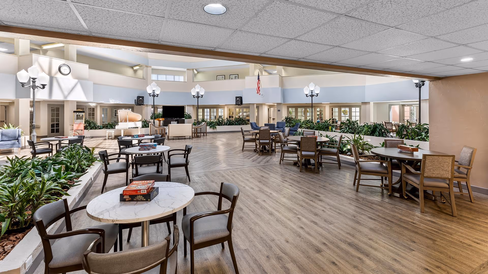 Spacious and well-lit common area in a senior living facility with multiple round tables and chairs arranged for socializing or activities. The room features indoor plants in raised planters, a large flat-screen TV, a piano, and decorative streetlamp-style lights. The flooring is wood-style, and the ceiling has recessed lighting. The space is open with large windows and doors leading to other rooms.