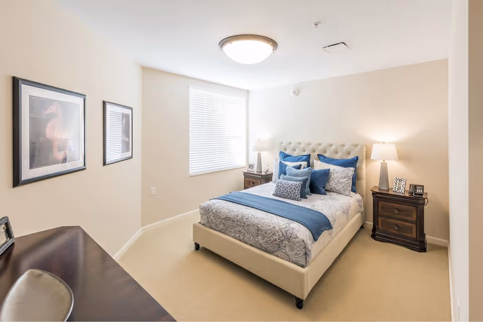 A bright bedroom with a beige upholstered bed featuring blue and patterned bedding and multiple pillows. There are two dark wooden nightstands on either side of the bed, each with a lamp and decorative items. A window with white blinds lets in natural light, and two framed pictures hang on the wall to the left. A dark wooden desk with a chair is partially visible in the foreground.