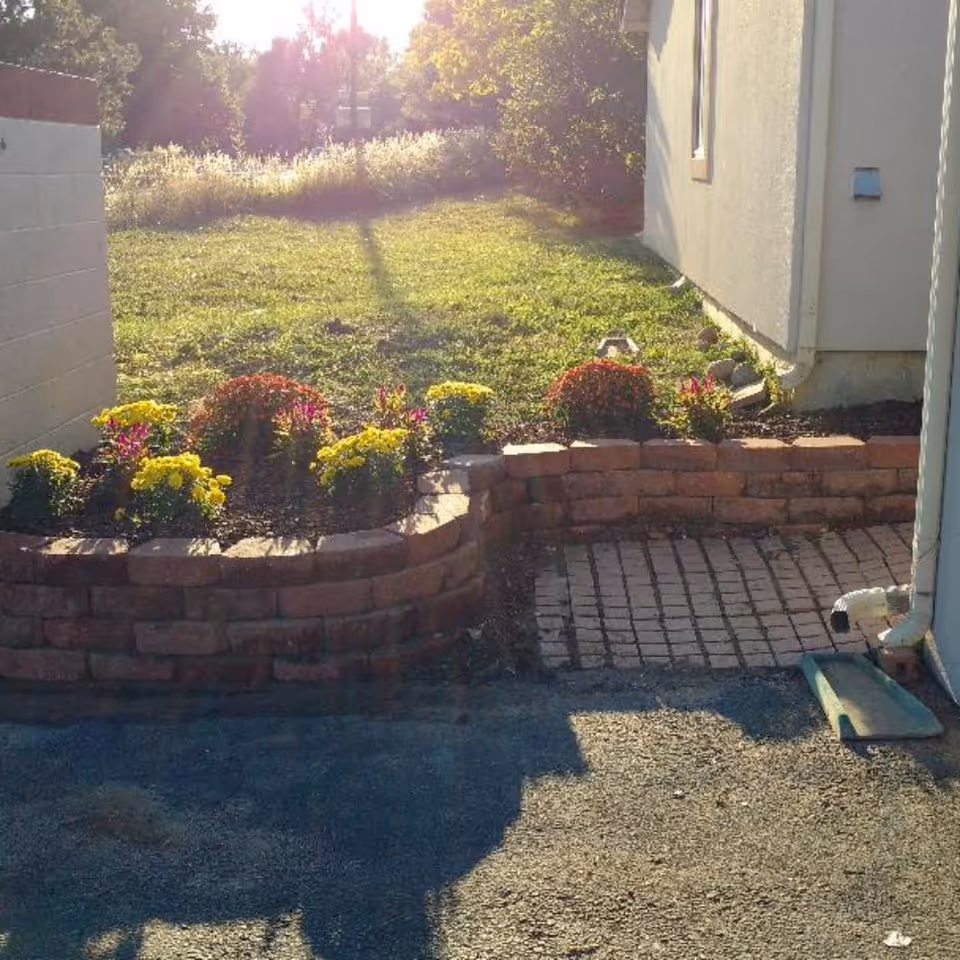 A small garden area with a raised brick flower bed containing yellow and red flowers, adjacent to a building wall with a window and a downspout. The garden is bordered by a paved pathway and a grassy lawn in the background with sunlight shining through trees.