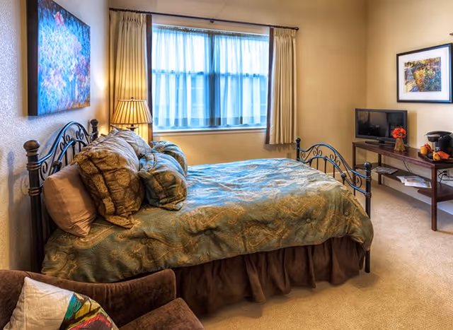 A cozy bedroom with a metal-framed bed covered in a patterned blue and brown bedspread and multiple pillows. There is a window with sheer and beige curtains letting in natural light. A bedside lamp is on a small table next to the bed. Across from the bed is a wooden TV stand with a small flat-screen TV, a coffee maker, and some decorative items. A framed picture hangs on the wall above the TV stand. In the foreground, part of a brown armchair with a colorful pillow is visible.
